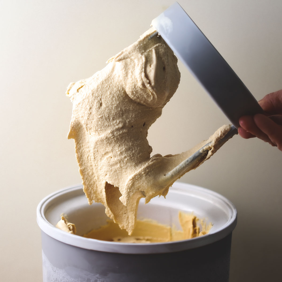An ice cream maker paddle just pulled out of the ice maker bowl with Caramel ice cream still attached to it; a hand is holding the paddle.