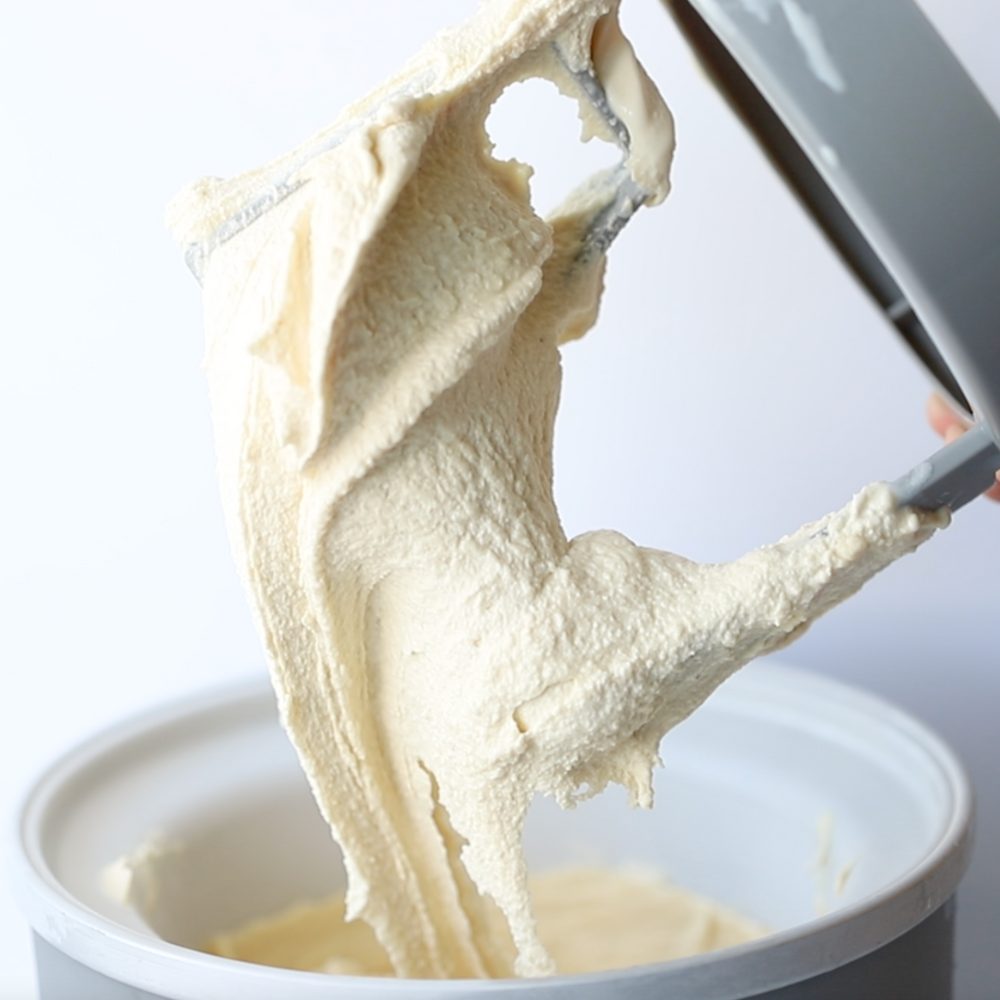 An ice cream maker paddle just pulled out of the ice maker bowl with coffee ice cream still attached to it; a hand is holding the paddle to show the stretchy texture of the icy cream.